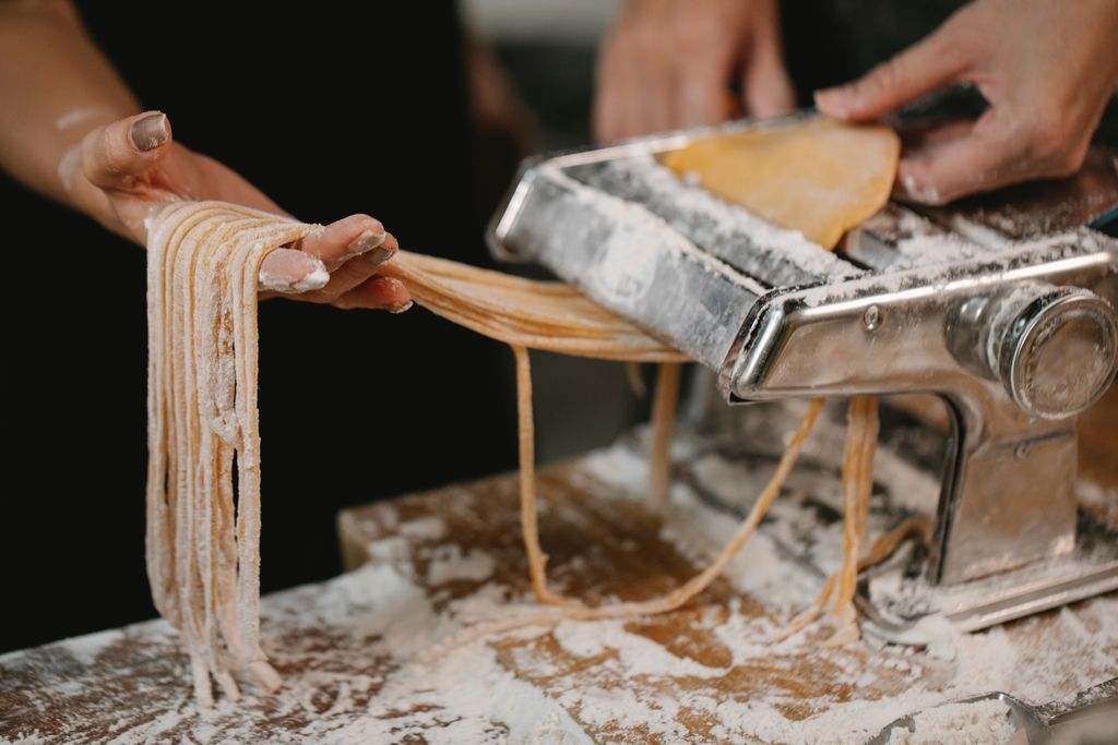 crop-anonymous-female-producing-long-noodle-with-metal-pasta-cutter-on-wooden-table-covered-with-flour